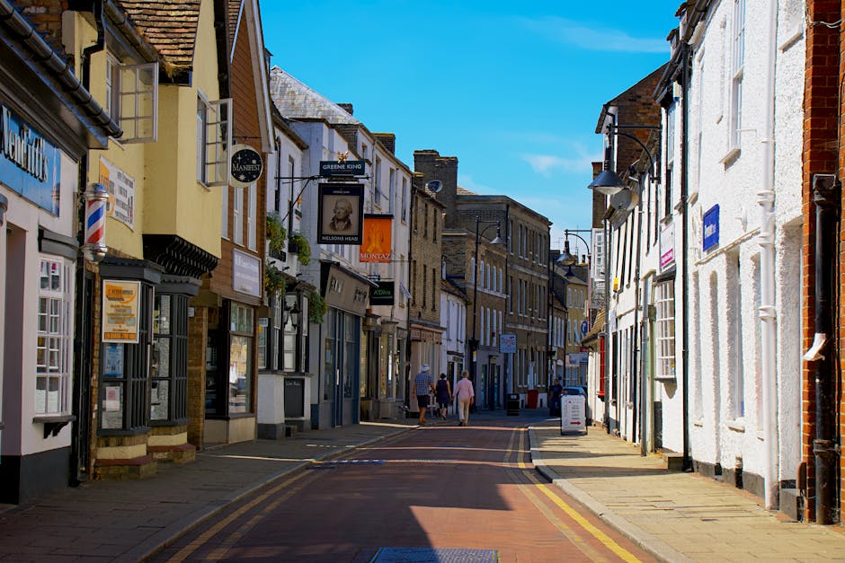 A narrow street in Wimbledon Village lined with small shops housed in historic buildings with various signboards, including barbershop, cafes, and boutiques. The shops have a mix of brick, stucco, and timber facades, some with hanging flower baskets and window displays. The street is paved with bricks and features double yellow lines along the curbs. A small white van is parked on the right side, partially visible, suggesting a home relocation or furniture transport process. Pedestrians are walking along the pavement, and there is outdoor lighting attached to the buildings. The sky is clear and blue, indicating daytime, and the scene reflects a typical busy high street scene that could be involved in packing, loading, or house removals, with the presence of vehicles and storefronts relevant to home relocation services in Wimbledon.