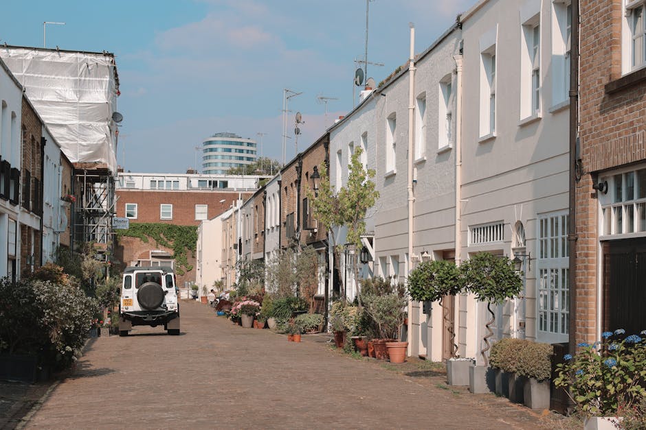 A residential street scene showing a white van parked on a narrow cobblestone pavement, used for home relocation purposes. The van is positioned at the center of the image, with its rear facing the viewer, and appears to be in the process of loading or unloading furniture and boxes. The surrounding area features terraced houses with white and brick facades, some with decorative plants and potted greenery placed along the pavement beside the entrances. Several cardboard boxes, plastic wrapping, and blankets are visible on the driveway or near the doorway, indicating packing materials used for furniture transport. The street is well-lit with natural daylight, and the environment appears calm, suitable for a professional moving or packing and moving service. In the background, a modern multi-storey building rises above the row of houses, emphasizing the urban setting typical for house removals in Wimbledon. The scene illustrates the straightforward logistics involved in house removals and the careful handling of belongings in a confined residential area, aligning with the services offered by Man and Van Wimbledon for narrow streets and home relocation tasks.