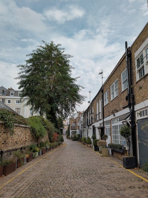 A narrow street in Wimbledon Village lined with small shops housed in historic buildings with various signboards, including barbershop, cafes, and boutiques. The shops have a mix of brick, stucco, and timber facades, some with hanging flower baskets and window displays. The street is paved with bricks and features double yellow lines along the curbs. A small white van is parked on the right side, partially visible, suggesting a home relocation or furniture transport process. Pedestrians are walking along the pavement, and there is outdoor lighting attached to the buildings. The sky is clear and blue, indicating daytime, and the scene reflects a typical busy high street scene that could be involved in packing, loading, or house removals, with the presence of vehicles and storefronts relevant to home relocation services in Wimbledon.