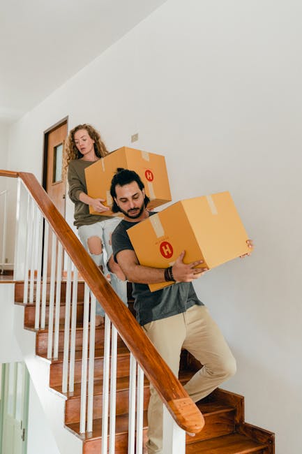 A young woman with dark brown hair, wearing a beige jacket, is seen carrying a large cardboard box up a staircase as part of a home relocation process. Behind her, a man with short hair, dressed in a